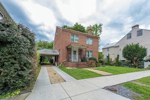 A two-story brick house with a covered porch, a front yard with a pathway, and a neighboring house. - Charming Shepherdstown Home - Walk to Downtown (Shepherdstown)