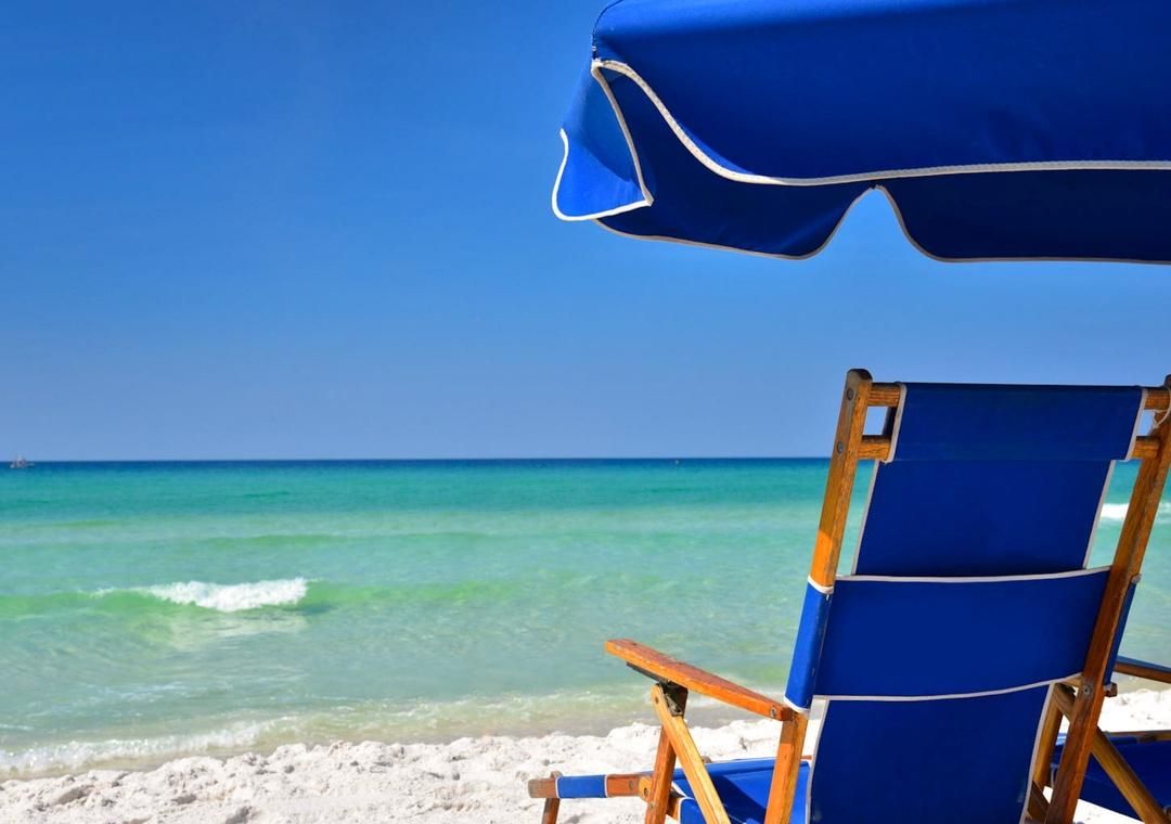 Escena de playa con cielo despejado, una silla de madera y una sombrilla azul.