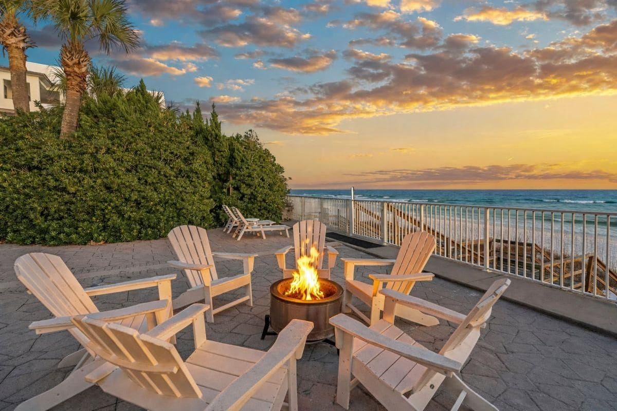 Een terras met Adirondack-stoelen rond een vuurkuil, met uitzicht op een strand en de oceaan.