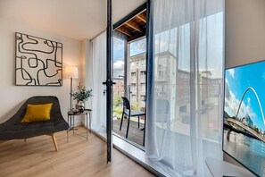 A modern living room with a sofa, a table with a lamp, and a large window with a view of buildings and a bridge. - Stephenson View (Gateshead)