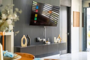 A modern living room with a flat-screen TV mounted on a wall with vertical lines, a console table with decorative items, and a door leading to another room. - Sea La Vie | Pool |2BR | Gated | Walk to the Beach (Llandovery)