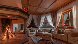A cozy living room with a fireplace, patterned sofas, and large windows with curtains.