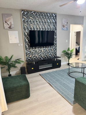 A modern living room with a flat-screen TV mounted on a geometric-patterned wall, a glass coffee table, and two green ottomans. - ABBY COURT (Mandeville)