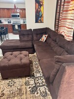 Una sala de estar con un sofá seccional y un reposapiés, una alfombra estampada, y una cocina al fondo.