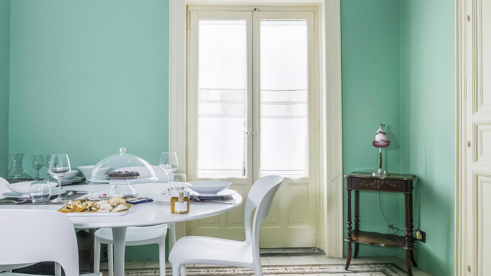 Un coin repas avec une table ronde dressée pour quatre personnes, des chaises blanches et une fenêtre équipée de stores.