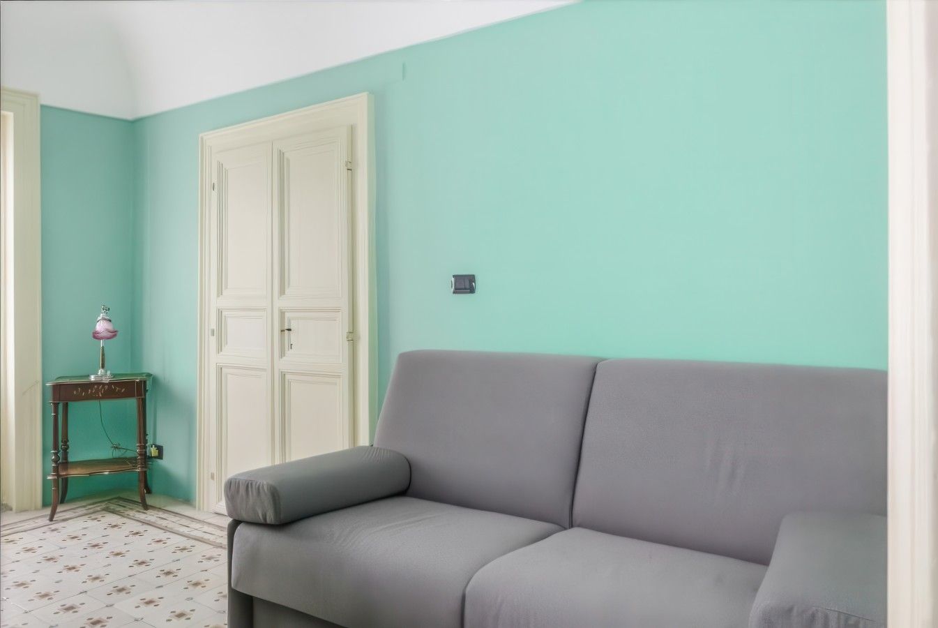 Une pièce avec un canapé gris, une petite table avec une lampe et une porte blanche.