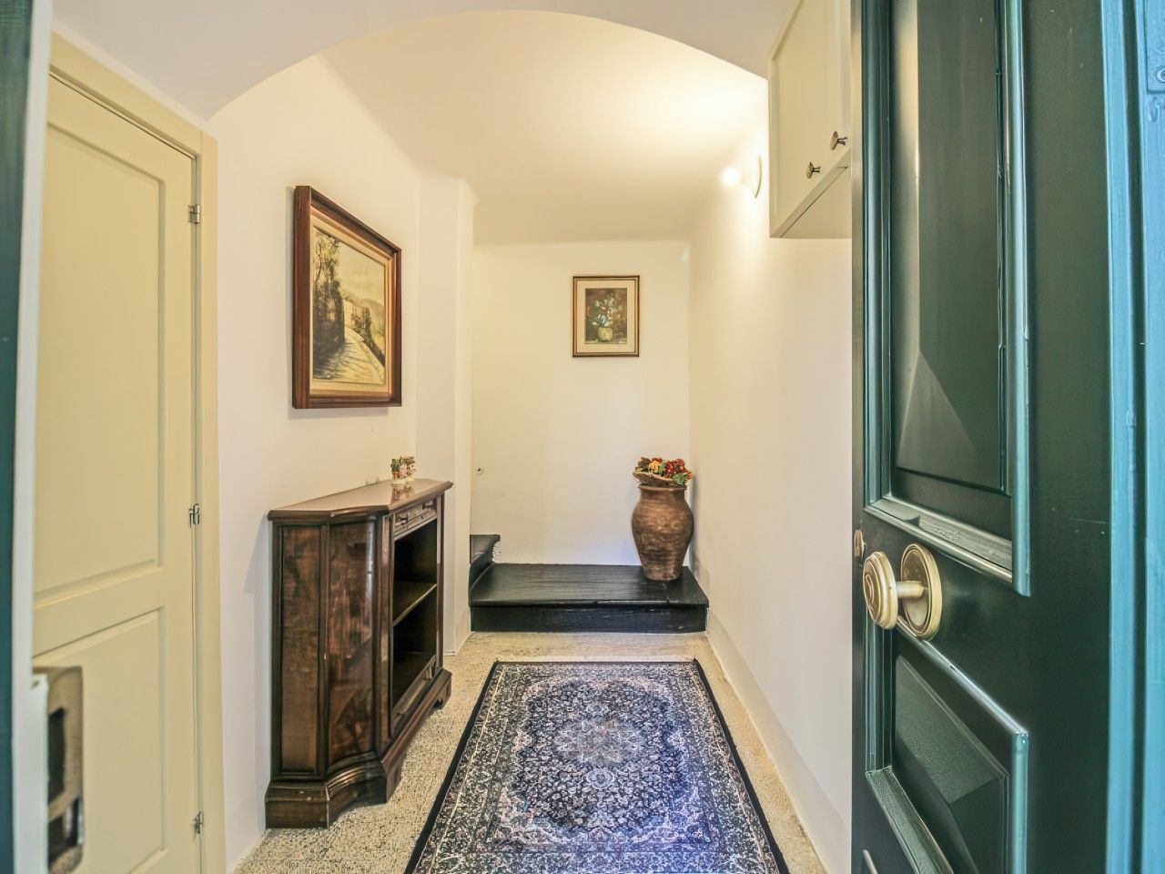 Un corridoio con una porta scura, un piccolo ripiano con oggetti decorativi, un tappeto e due quadri appesi al muro.