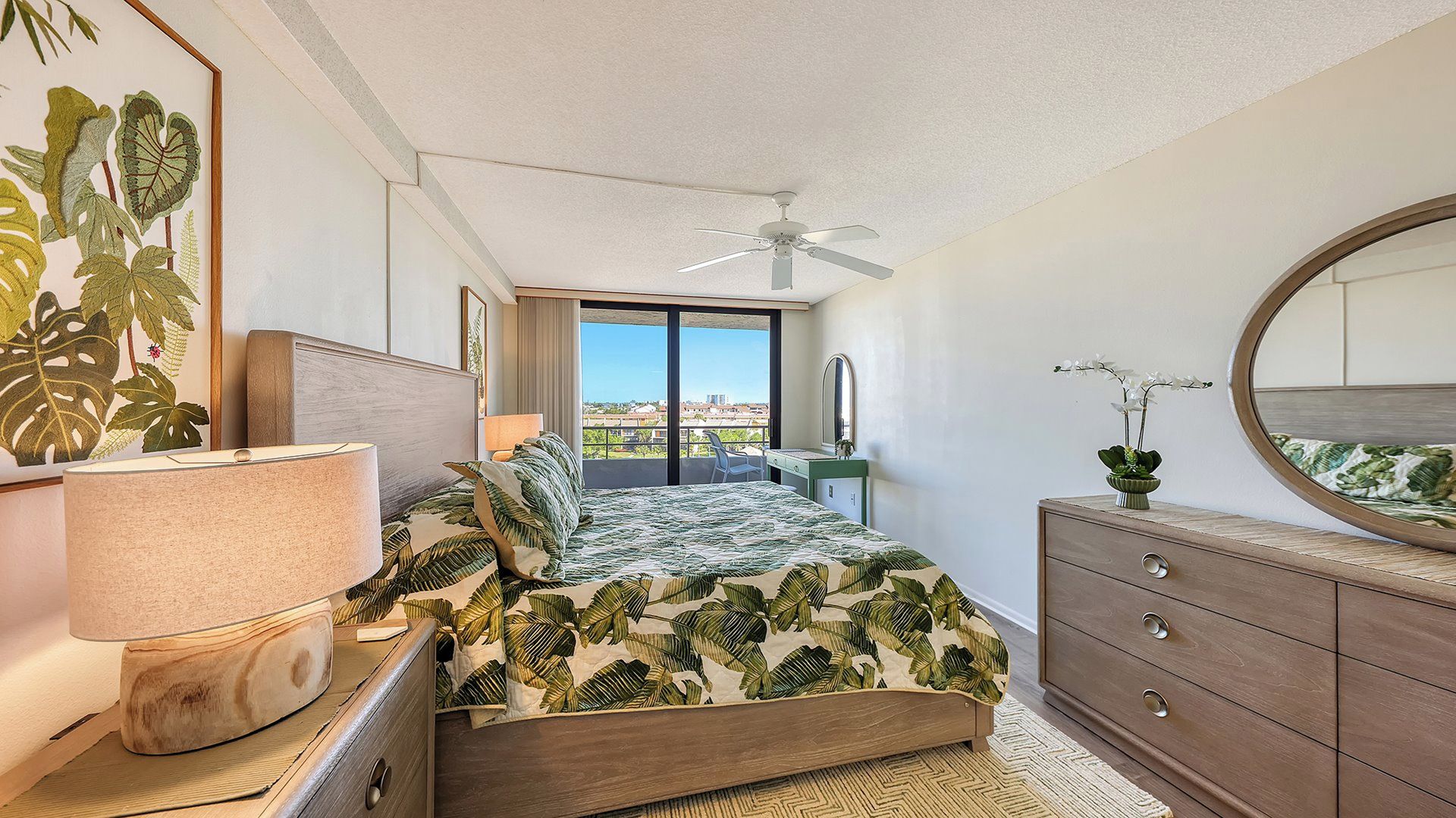 Une chambre à coucher comprenant un lit, une commode, un miroir, un ventilateur de plafond et une vue sur la ville grâce à de grandes fenêtres.