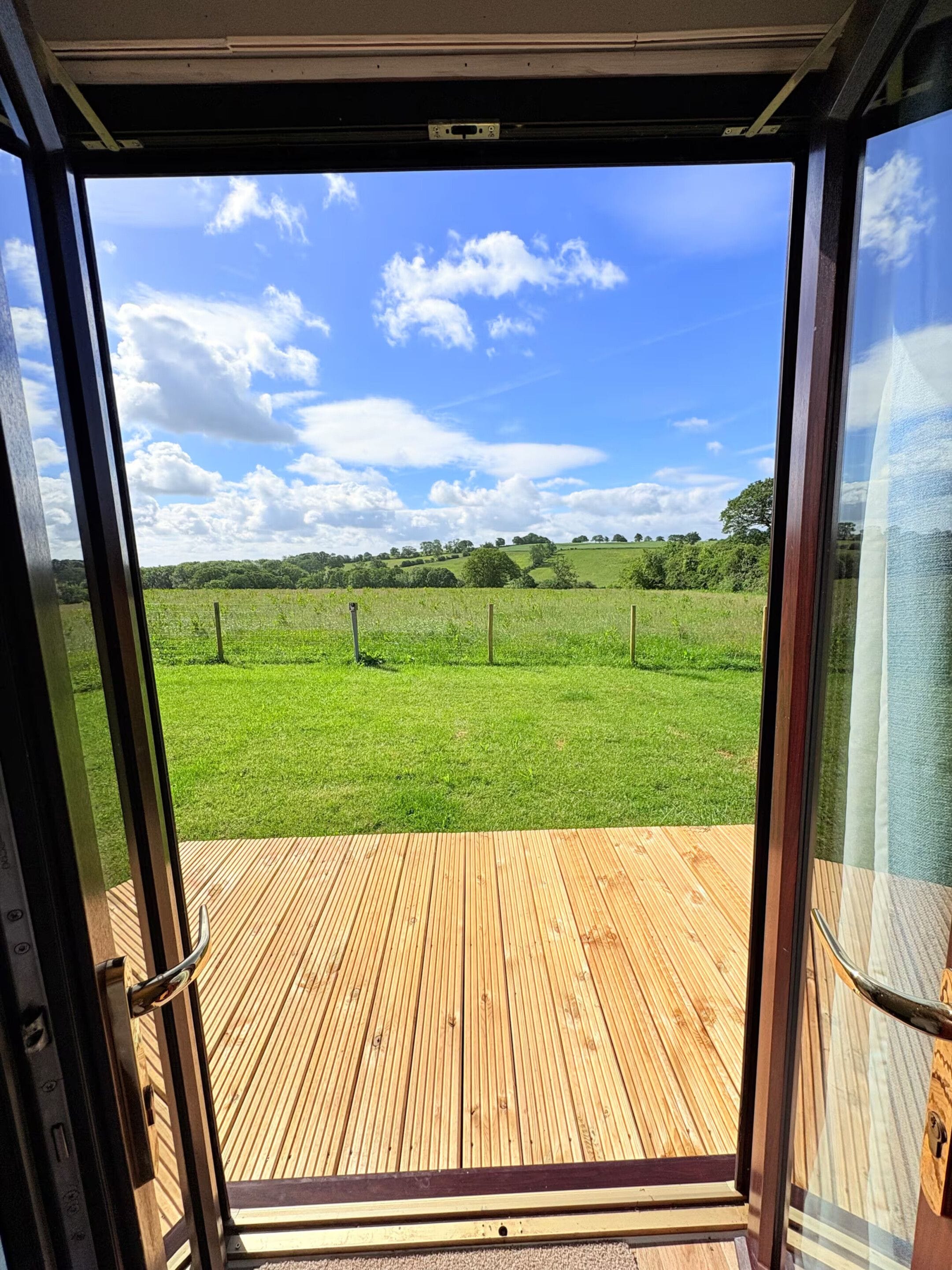 Puerta abierta que da a una terraza de madera con vistas a un campo verde y cielo despejado.