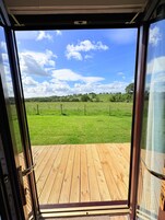 Puerta abierta que da a una terraza de madera con vistas a un campo verde y cielo despejado.