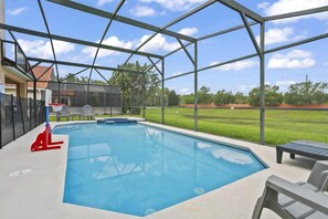 Piscina com cesta de basquete e cadeiras, cercada por uma área coberta com vista para um campo gramado.