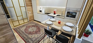 Eine moderne Küche mit weißen Schränken, einer Mikrowelle und einem Backofen. Ein Esstisch ist gedeckt mit Tellern, Gläsern und einer Kerze. Auf dem Boden liegt ein gemustertes Teppich, und Stühle stehen um den Tisch.