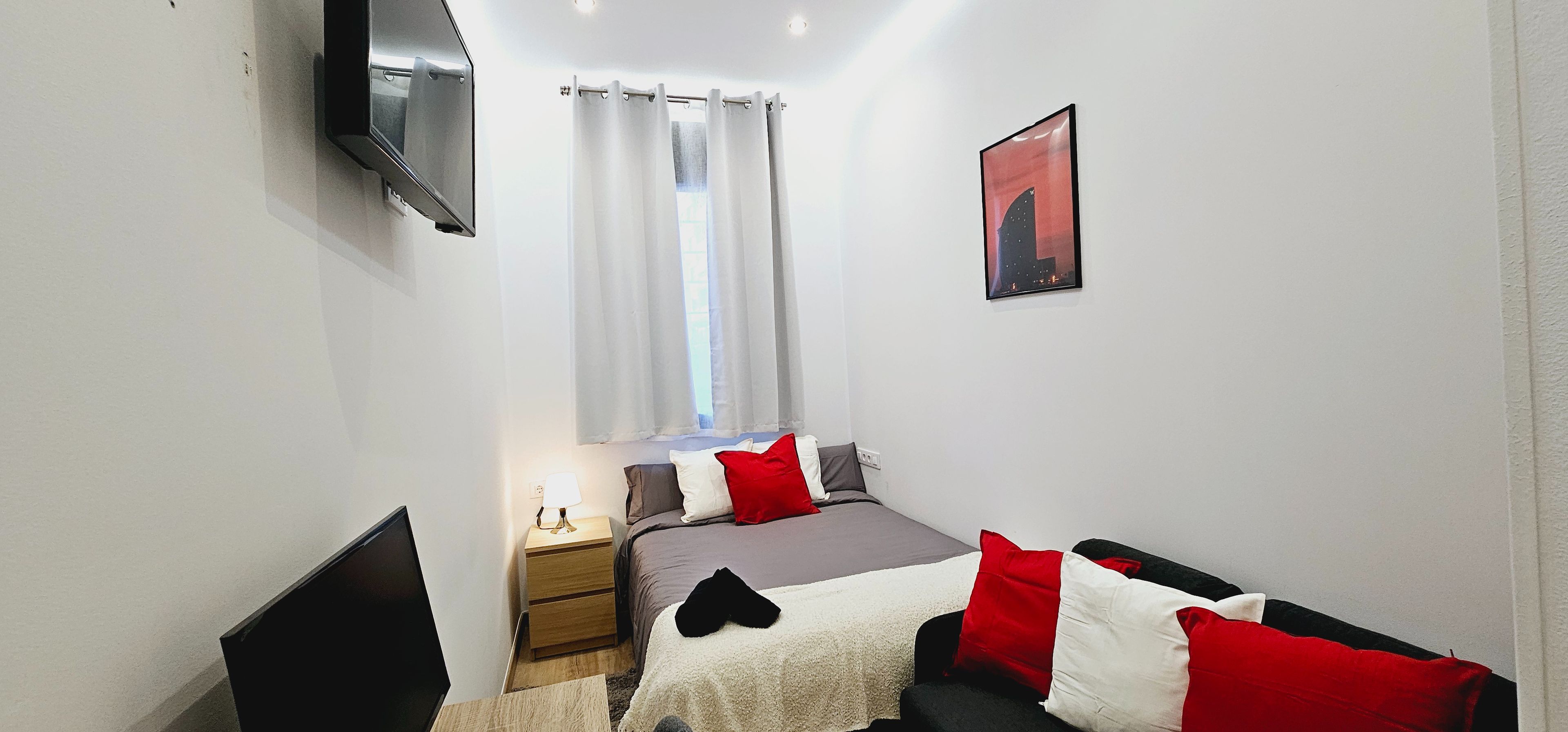 Un dormitorio con cama, mesita de noche, lámpara y televisor montado en la pared.