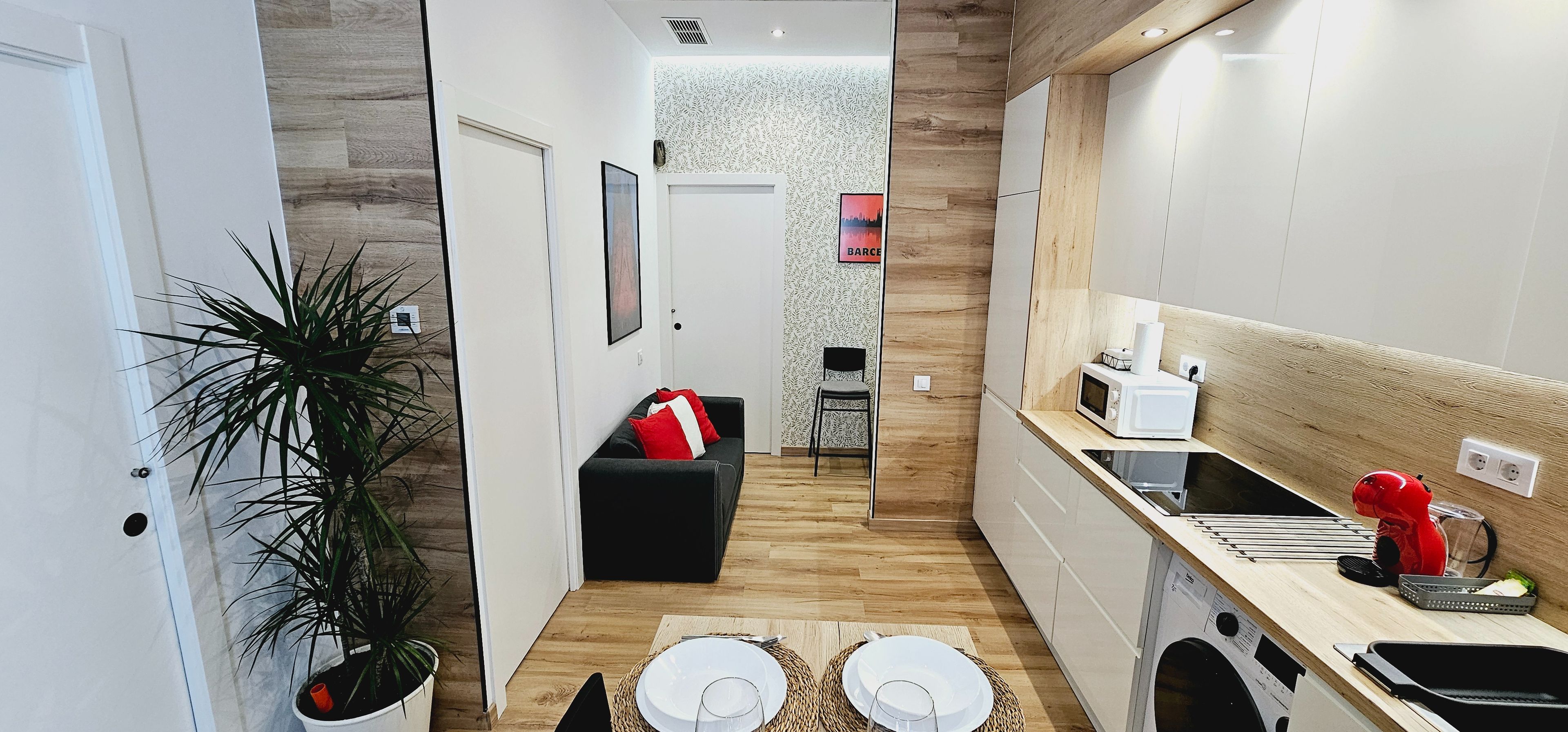 Una cocina moderna con armarios blancos, un microondas y un lavamanos. Hay una zona de comedor con una mesa preparada para dos personas, y un sofá negro con cojines rojos.