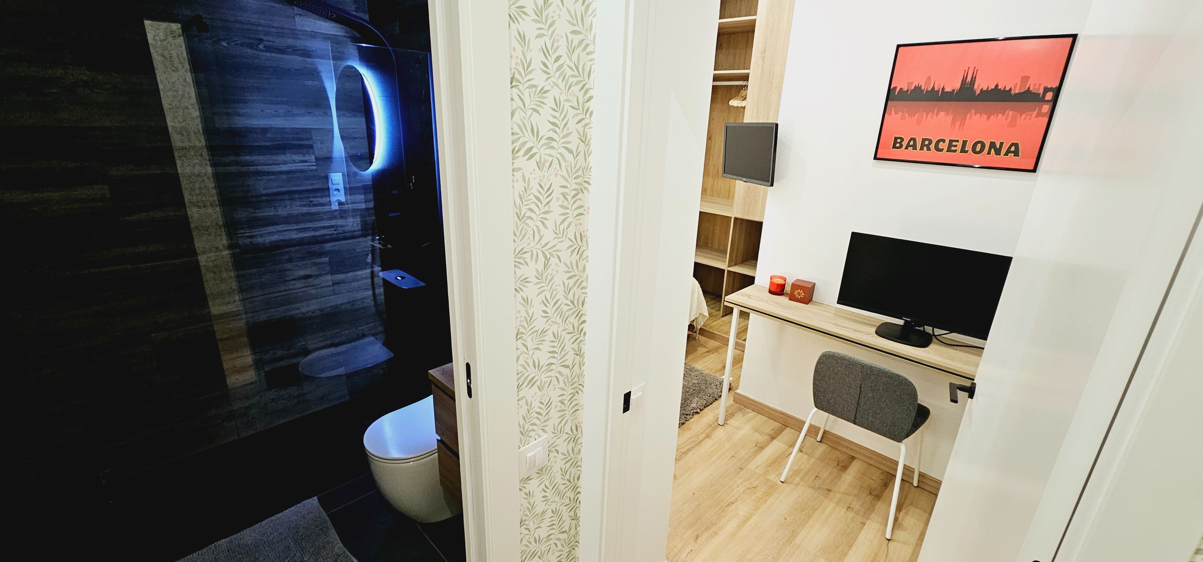 Ett modernt badrum med en duschkabin i glas och en toalett. Det finns ett litet skrivbord med en dator och en stol, och en inramad tavla med ett motiv från Barcelona hänger på väggen.