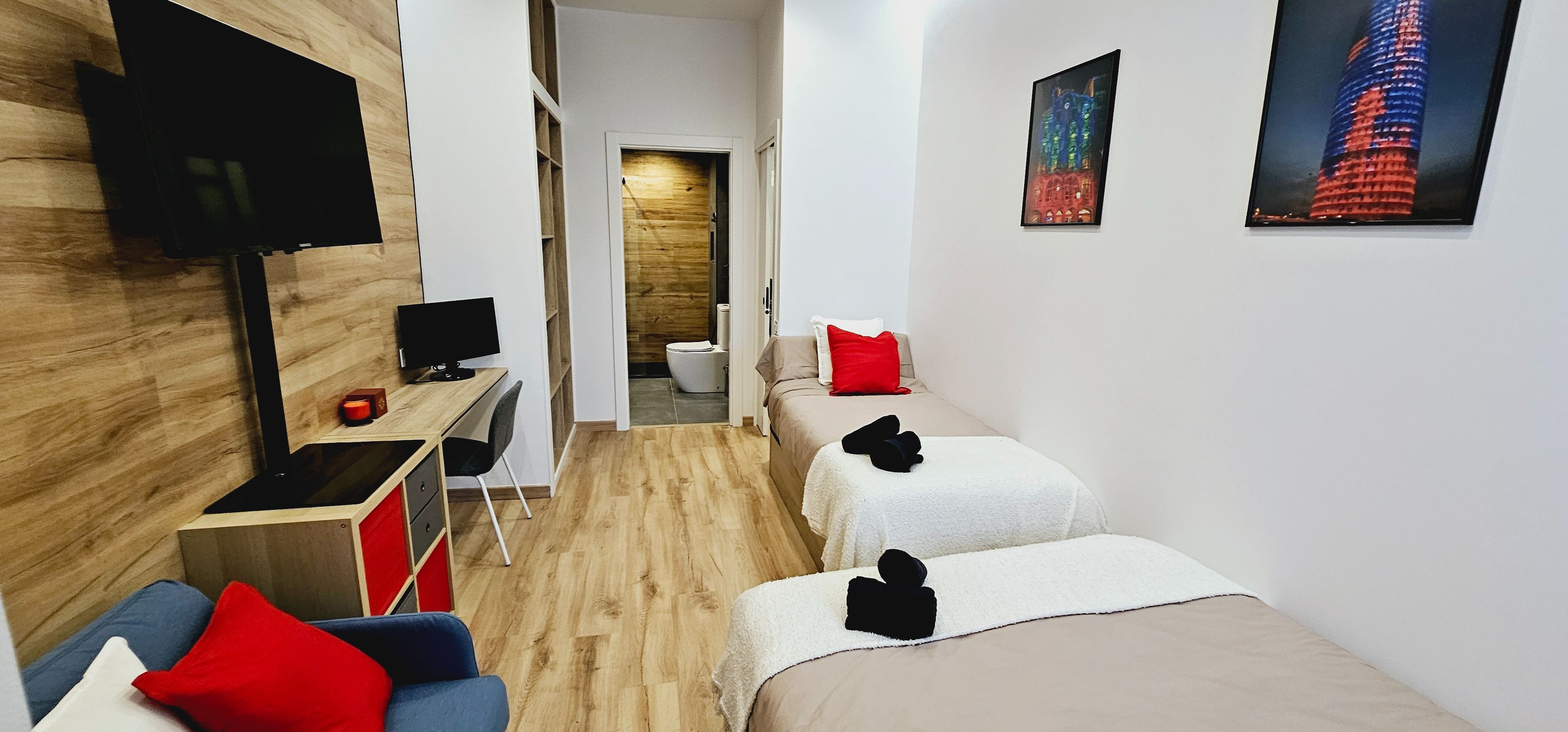 A modern hotel room with a wooden floor, a desk with a computer, a TV, a bed with white bedding, and a red pillow.