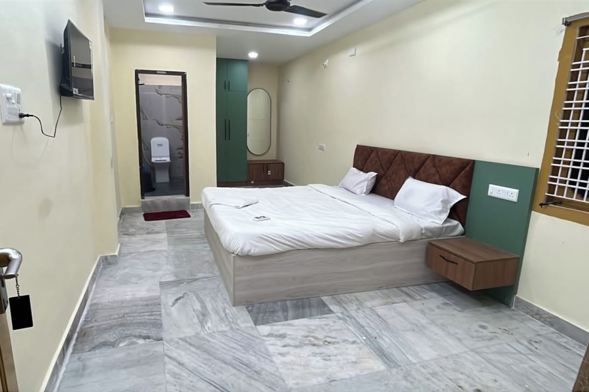 Une chambre à coucher comprenant un lit, des tables de chevet, un ventilateur de plafond, ainsi qu’une salle de bain avec des toilettes et un miroir.