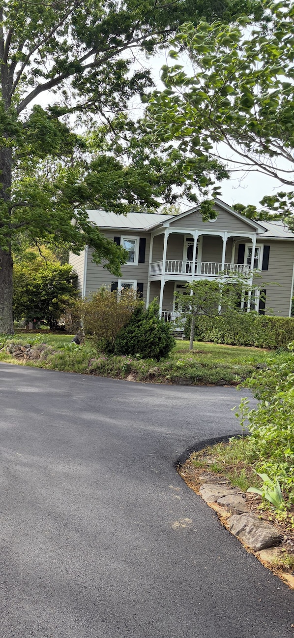 A two-story house with a front porch, surrounded by greenery and a paved driveway.