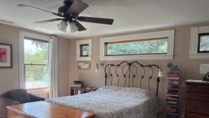 Ein Schlafzimmer mit Bett, Kommode, Nachttisch und einem Fenster mit Blick auf Bäume.