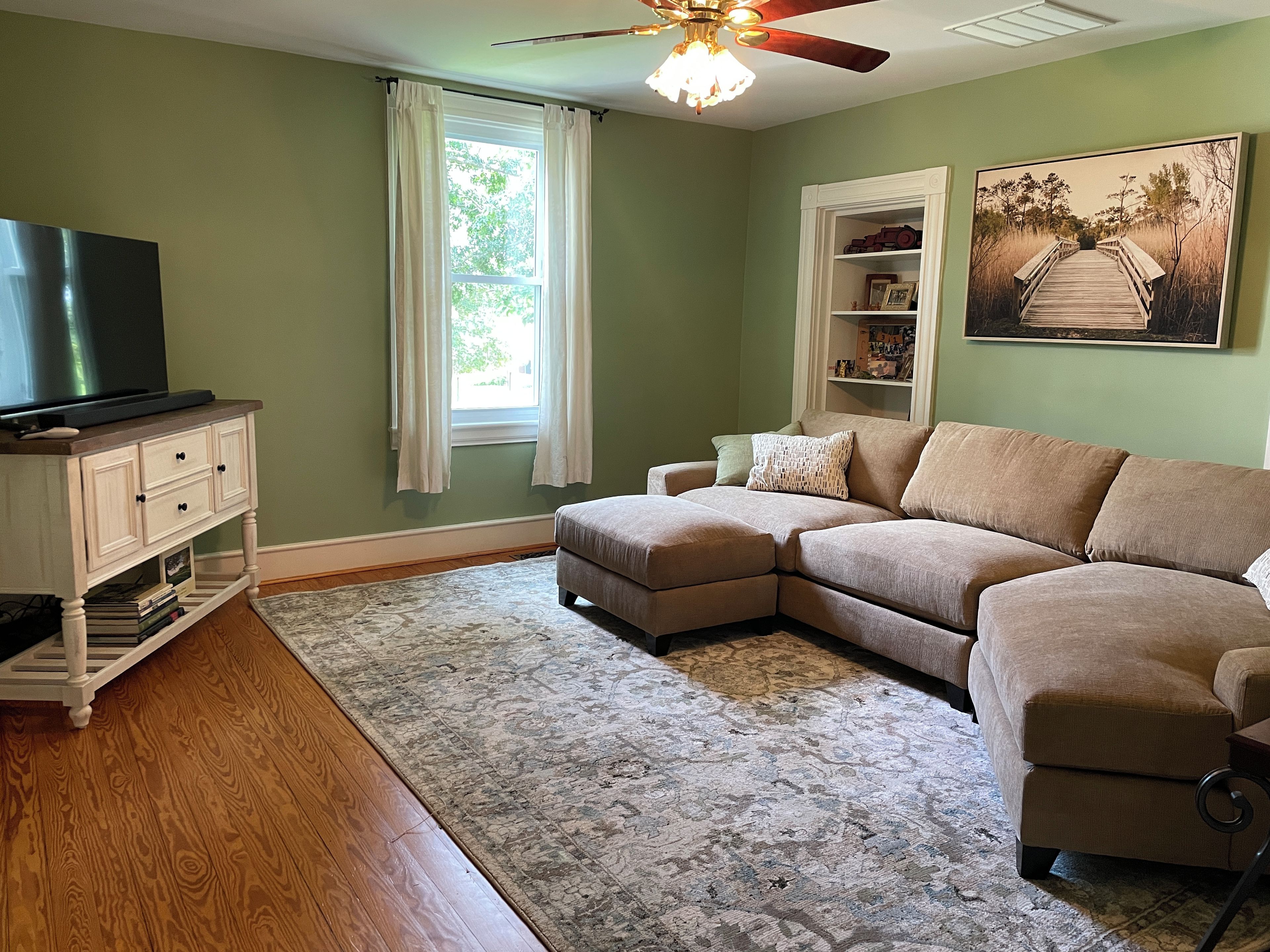 A living room with a sectional sofa, a flat-screen TV on a cabinet, a ceiling fan, and a large window with curtains.