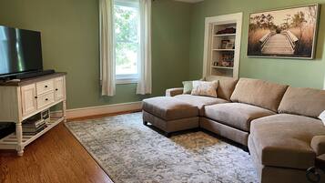A living room with a sectional sofa, a flat-screen TV on a cabinet, a ceiling fan, and a large window with curtains.