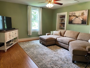 A living room with a sectional sofa, a flat-screen TV on a cabinet, a ceiling fan, and a large window with curtains.