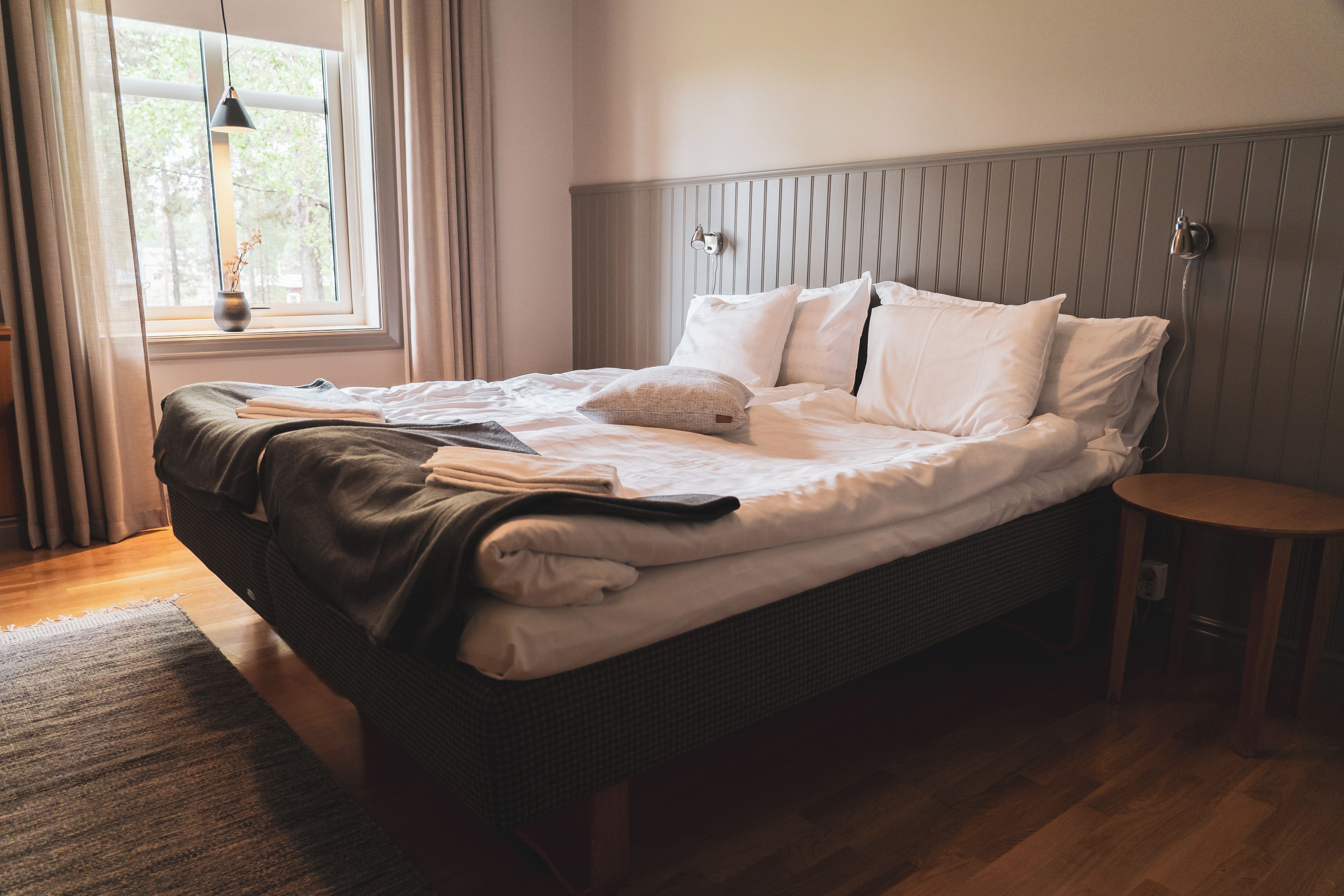 A neatly made bed with pillows and a blanket, a bedside table, a window with curtains, and a wall-mounted light fixture.