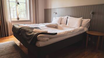A neatly made bed with pillows and a blanket, a bedside table, a window with curtains, and a wall-mounted light fixture.