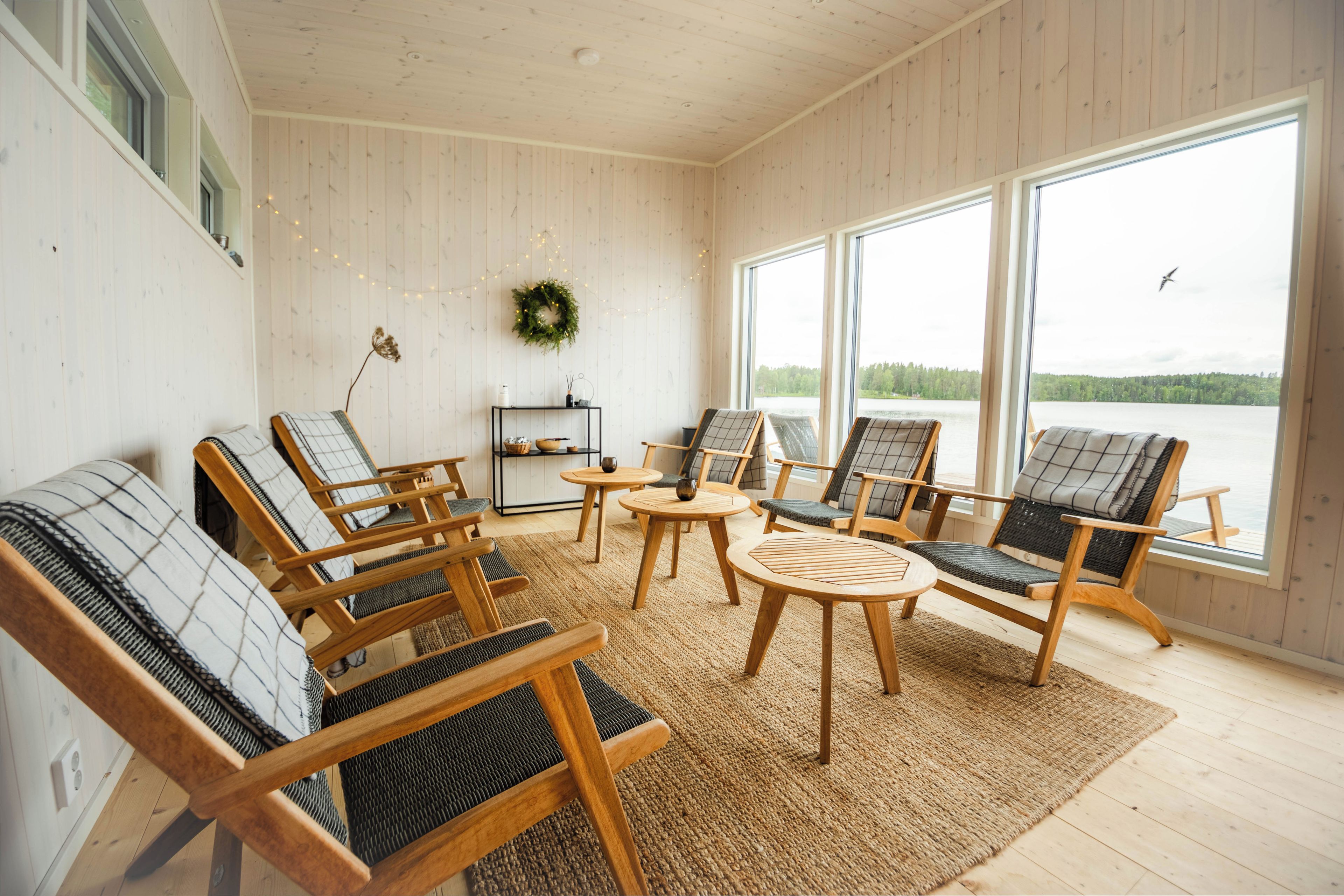 Ett mysigt rum med trägolv, ett stort fönster med utsikt över en sjö och flera sittgrupper med kuddar och ett litet bord.