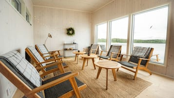 Ett mysigt rum med trägolv, ett stort fönster med utsikt över en sjö och flera sittgrupper med kuddar och ett litet bord.