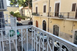 Ein Balkon mit weißen schmiedeeisernen Geländern, der auf eine Straße mit geparkten Autos und ein Gebäude mit Rundbögen blickt.