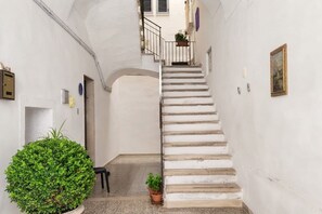Ein schmaler Gang mit einer weißen Treppe, die zu einer oberen Etage führt. Auf der linken Seite befinden sich Blumentöpfe und eine Tür.