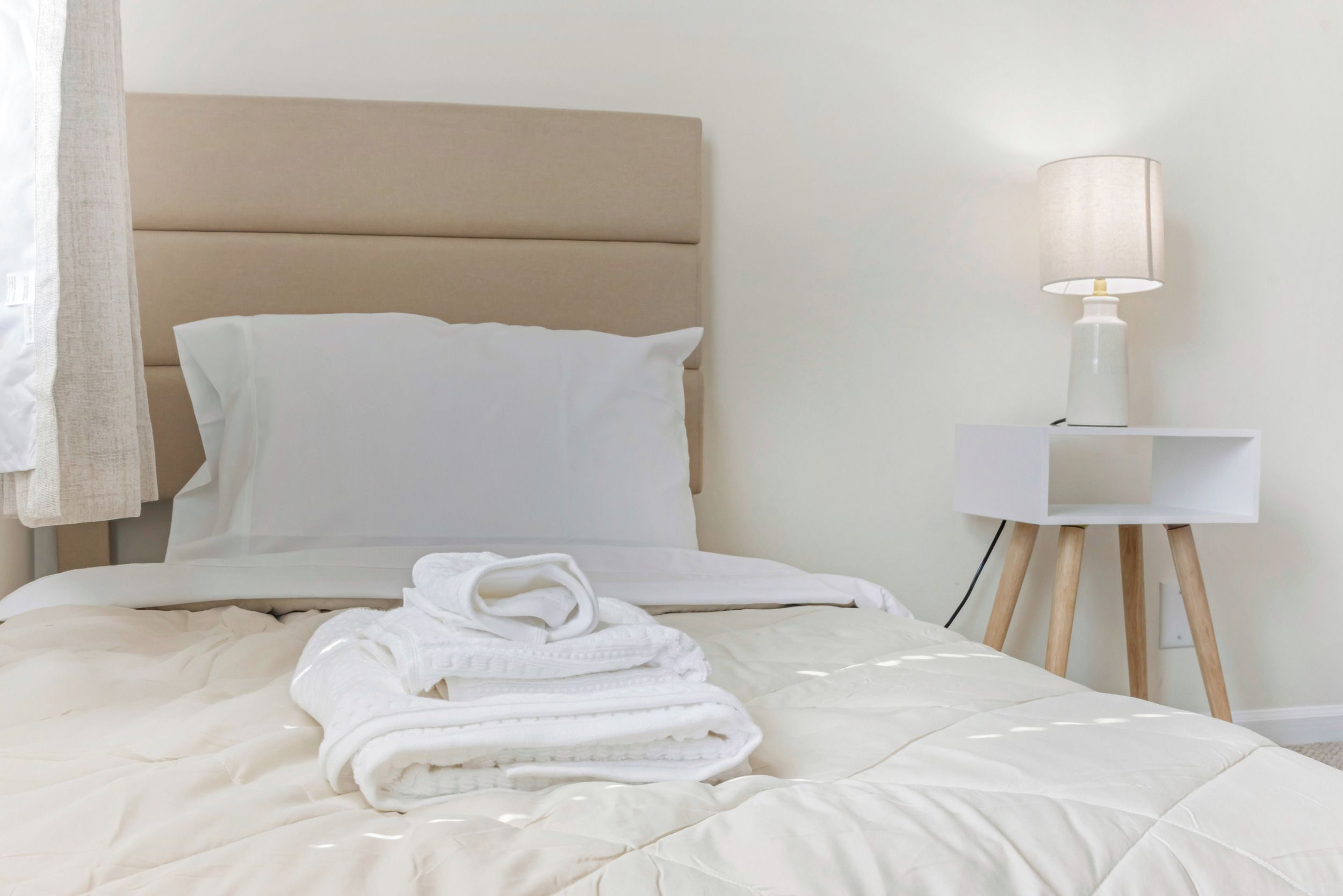 A neatly made bed with a beige headboard, a white bedside table with a lamp, and a rolled-up towel on the bed.