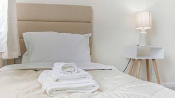 A neatly made bed with a beige headboard, a white bedside table with a lamp, and a rolled-up towel on the bed.