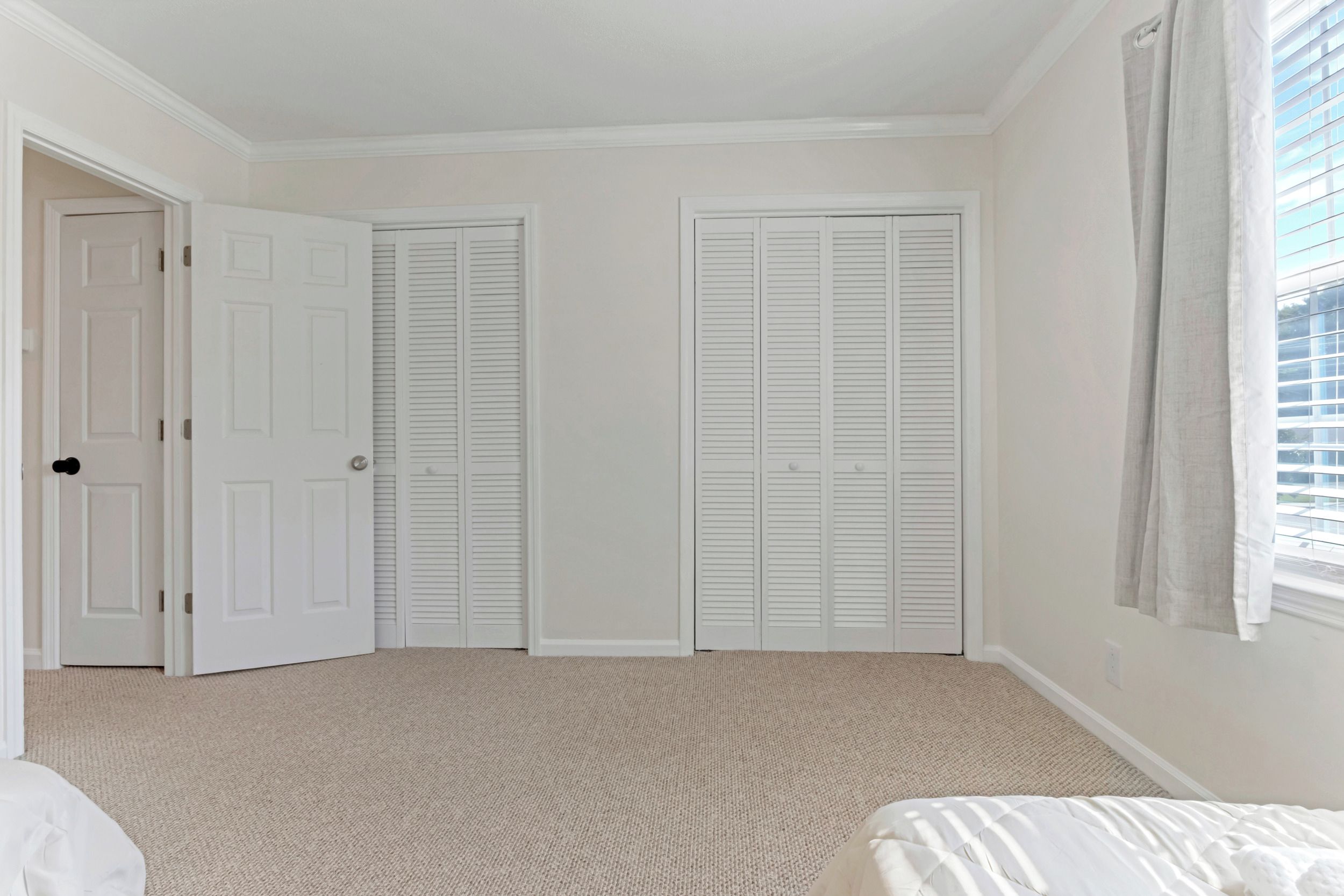 A bedroom with a carpeted floor, a bed, two white closets, and a window with blinds.