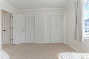 Ein Schlafzimmer mit Teppichboden, einem Bett, zwei weißen Schränken und einem Fenster mit Jalousien.