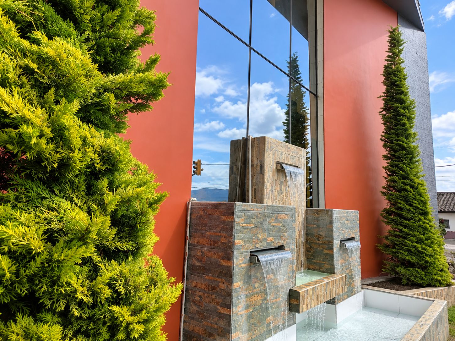 A modern building with a red facade, a stone water feature, and a tall coniferous tree.