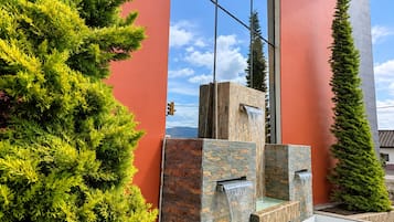 Um edifício moderno com fachada vermelha, um elemento de água em pedra e uma alta conífera.