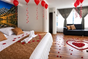 A hotel room with a bed, a sofa, and a heart-shaped arrangement of rose petals on the floor.