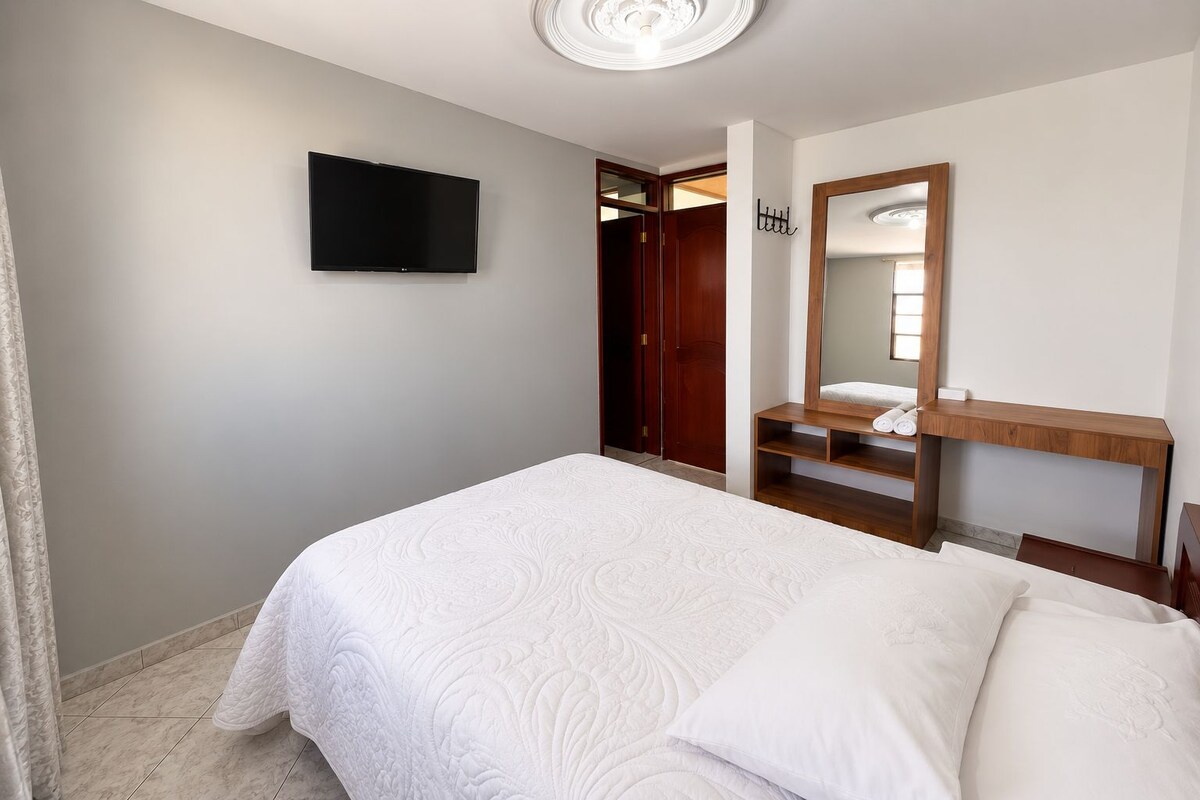 A hotel room with a bed, a television mounted on the wall, a wooden vanity with a mirror, and a door leading to another room.