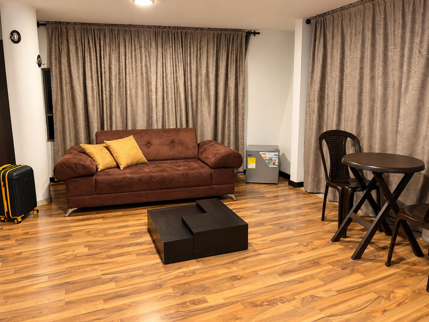 A furnished room with a sofa, a small table, a chair, a suitcase, and a refrigerator.