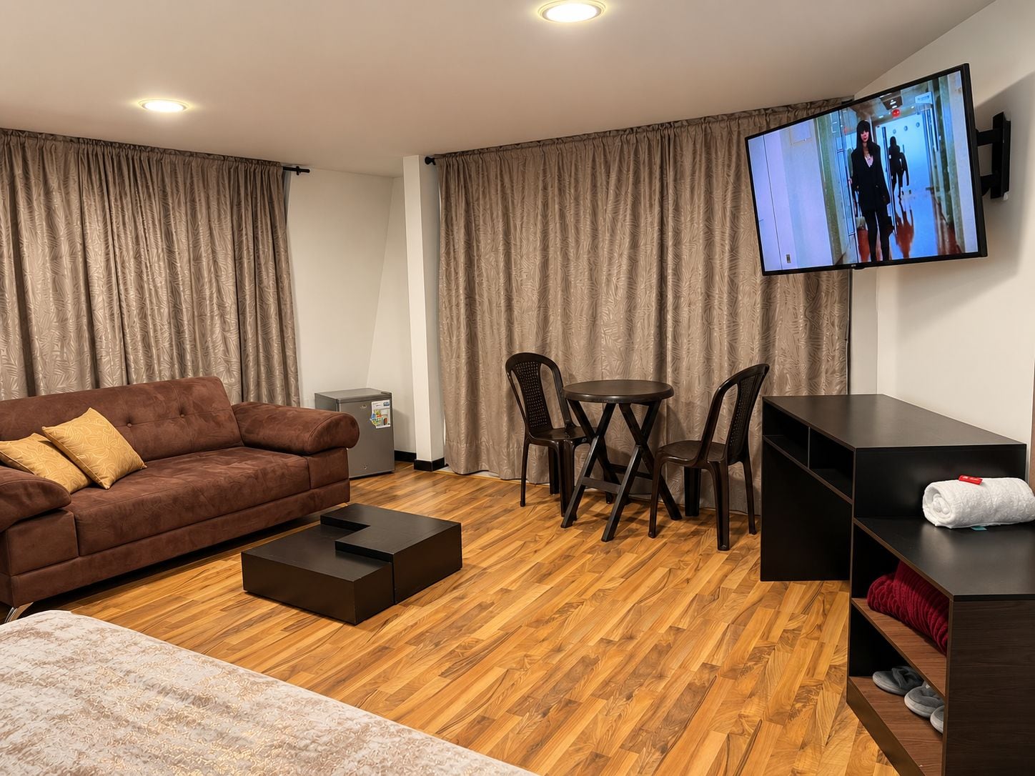 A modern living room with a brown sofa, a small table, and chairs. A television is mounted on the wall, and there is a wooden floor.