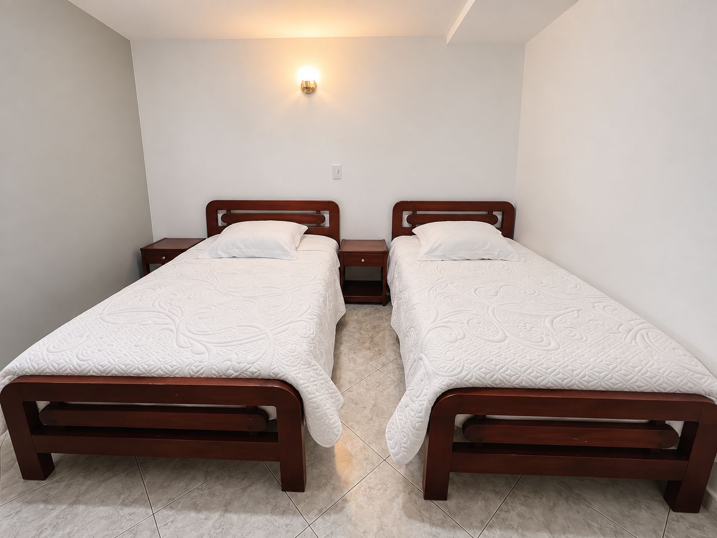 A room with two wooden beds, each with a white bedspread and pillows. There is a wall-mounted light fixture above the beds.