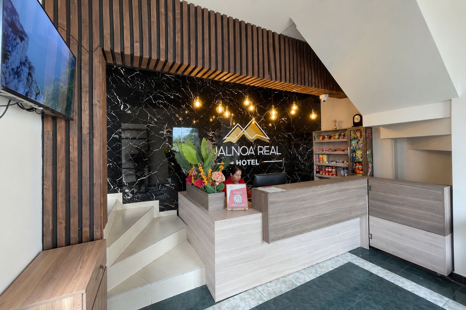 Un comptoir d’accueil d’hôtel au design moderne, avec un mur en marbre et une enseigne indiquant « ALINOA REAL HOTEL ».