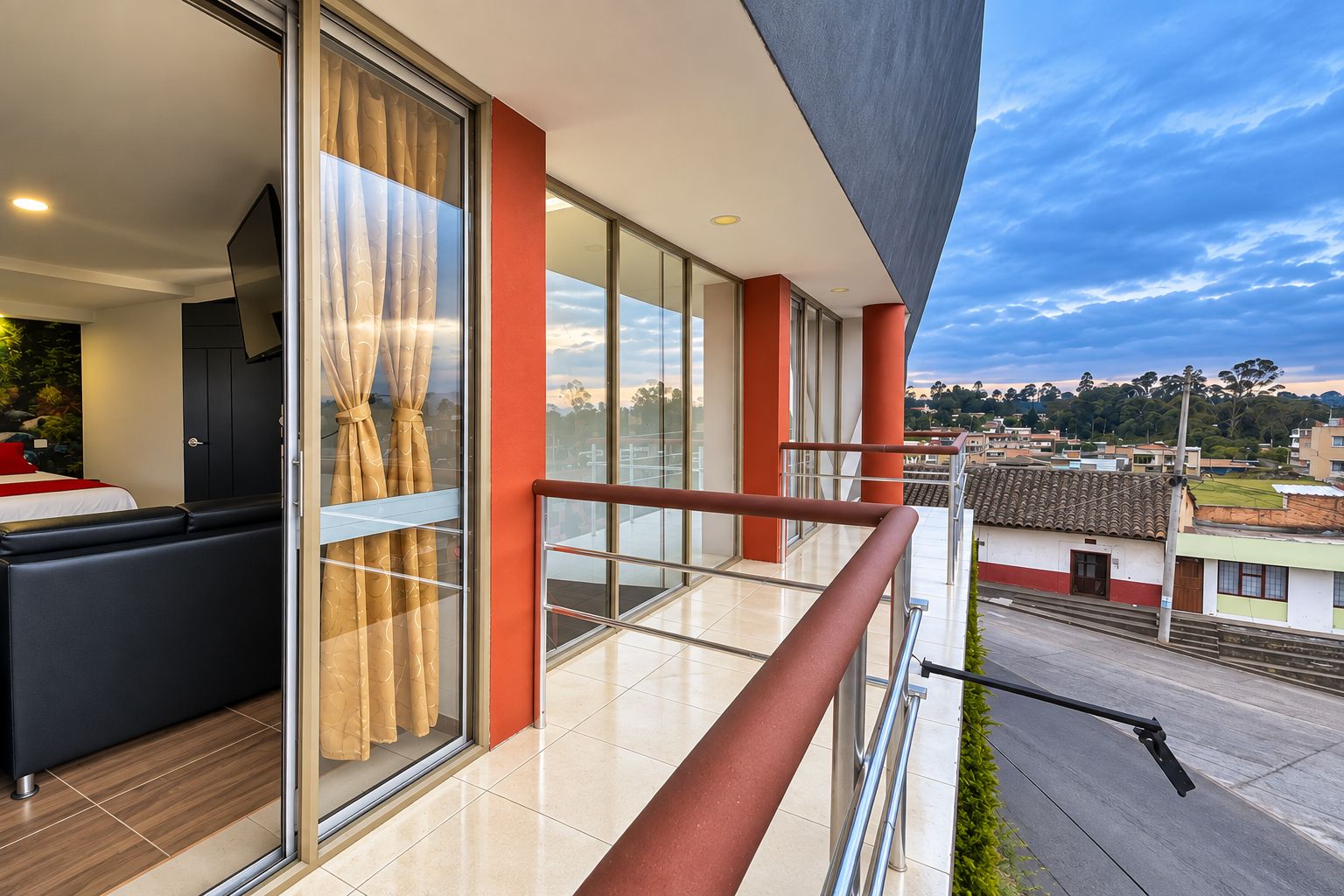A modern hotel room with a balcony, large windows, and a view of the cityscape.