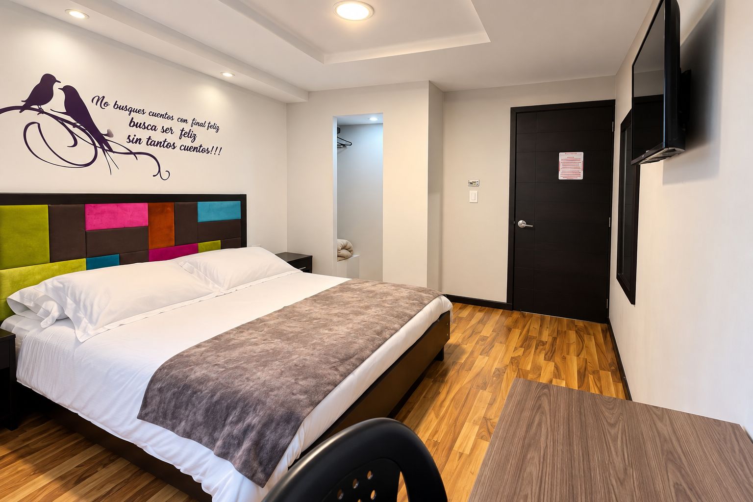 A hotel room with a bed, a desk, a chair, a wall-mounted TV, and a door.
