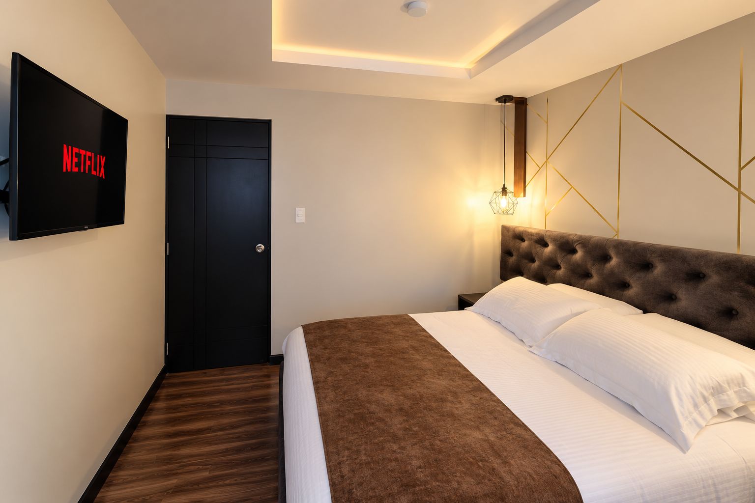 Habitación de hotel con una cama, televisor montado en la pared, una puerta y una lámpara también fijada a la pared.