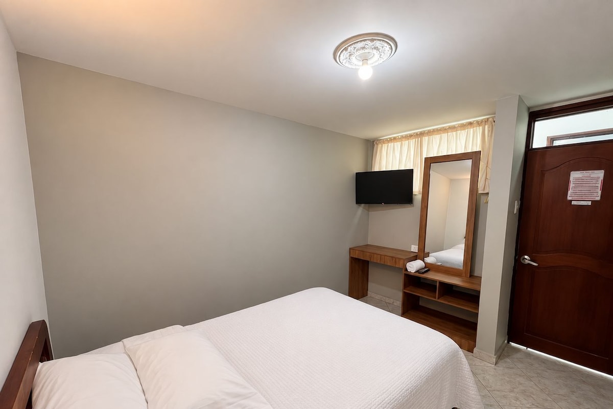 Una habitación de hotel con una cama, una mesita de noche de madera, un espejo, un televisor y una puerta.