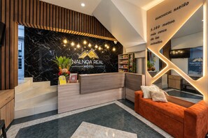 A hotel reception area with a marble wall, a reception desk, a sofa, and a staircase.