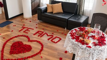 A room with a heart-shaped arrangement of rose petals spelling 'TE AMO' on the floor, a table with rose petals, strawberries, and nuts, and a black leather sofa with a pillow.